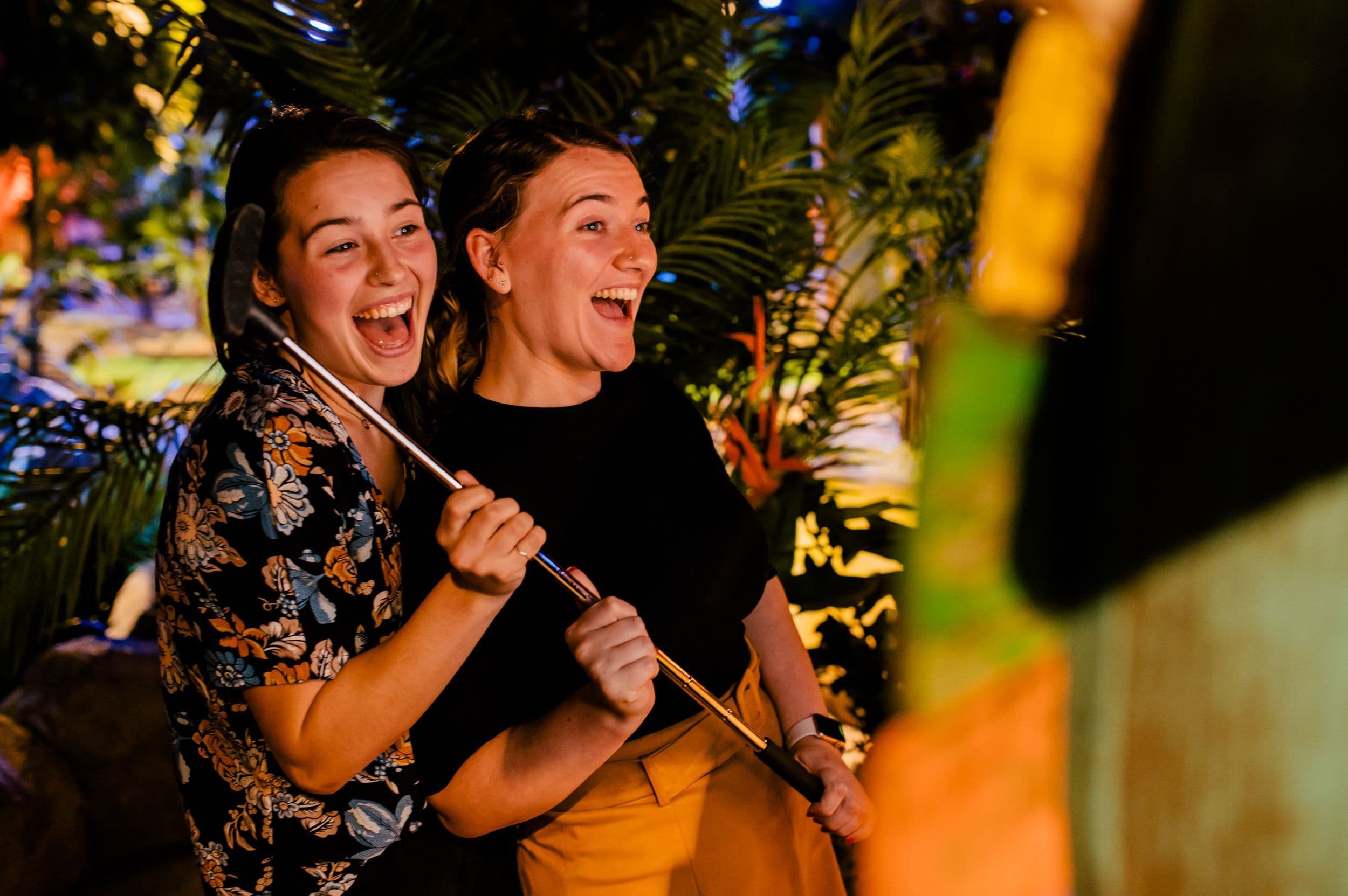 An image of two young women with their hair in ponytails smiling and holding their clubs up to the photobooth camera in orange and green light.